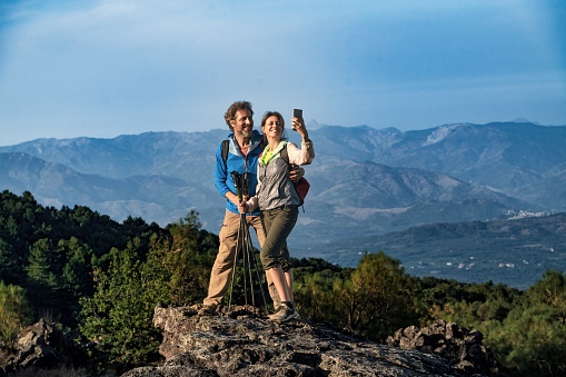 A middle-aged couple, in their 40s and 50s, stands atop a rocky peak with trekking gear, capturing a selfie with the breathtaking valley backdrop.
