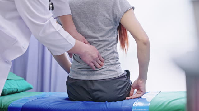 Rear view of a female patient sitting on a bed and having a lower back pain while a doctor touching her lower back to diagnose her condition in a doctor's office.