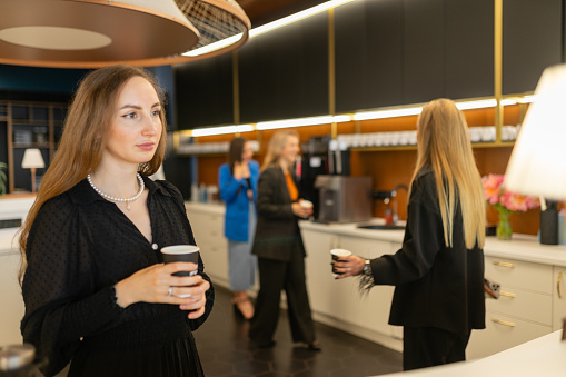 Contemplating woman holds cup with hot beverage in office kitchen. Female employees converse and enjoy pause from work in corporate kitchen