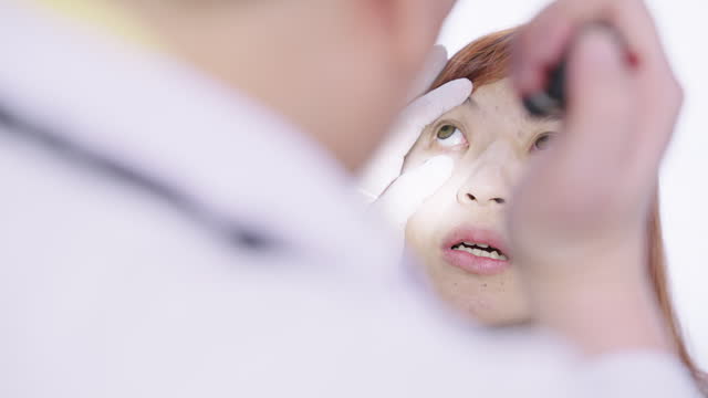 Headshot of a female patient looking up and rolling her eyes around while an optometrist examining her eyes condition by shining a flashlight on her eyes in a medical examination room.