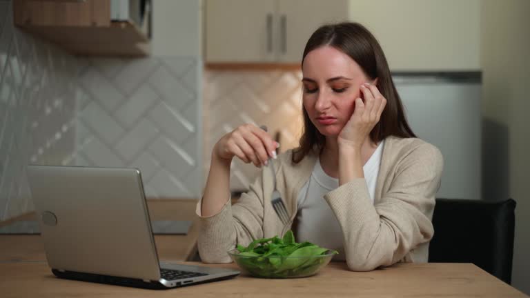 A thoughtful young woman eats a salad and experiences a lack of appetite while sitting in the kitchen and using a laptop. Digestive problems, as well as spoiled and tasteless food