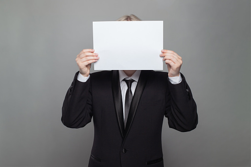 Anonymous businessman hiding his head behind blank empty white paper against gray studio wall background