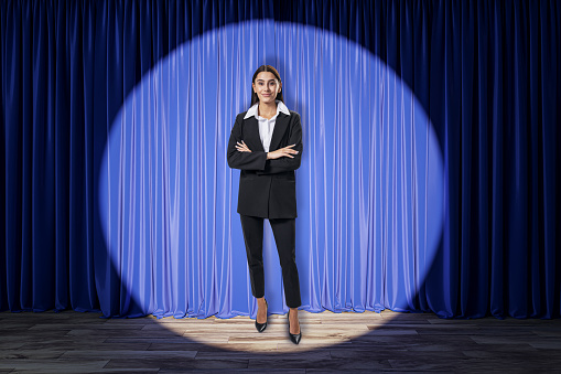 Attractive young businesswoman standing in front of blue curtains in spotlight in theater. Presentation and speech concept