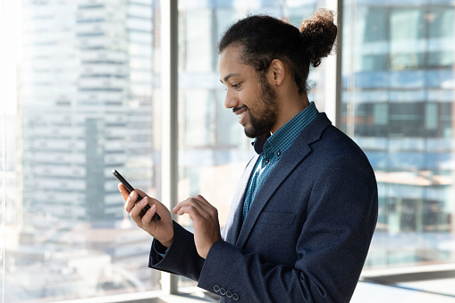 Side view smiling African American businessman using smartphone, standing near panoramic window, entrepreneur employee in suit typing on screen, writing message in social network, reading news