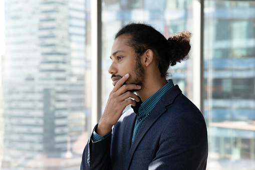 Head shot thoughtful African American businessman looking out window in office, pensive entrepreneur owner touching chin, pondering project strategy, thinking about business problem solution