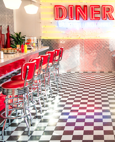 An old-fashioned American diner interior, with retro neon sign, red vinyl stools in a row by the counter, and a black and white chequered floor.