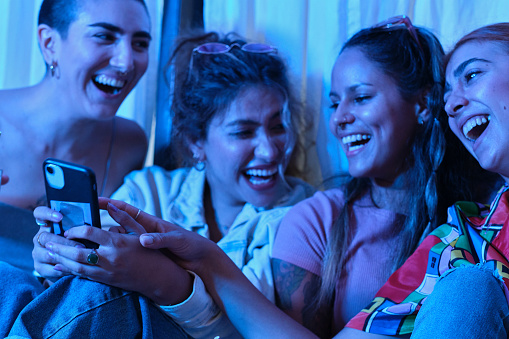 front view portrait of a group of generation z women with cool attitude and vintage outfits smiling and using an smartphone and joking