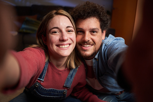 Happy young adult Caucasian couple taking smiling selfie looking at camera at home at night. Cheerful people in love posing mobile photo indoors. Millennial man and woman in living room at apartment.