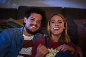 Young Caucasian smiling couple with popcorn watching movies TV on living room floor in evening.