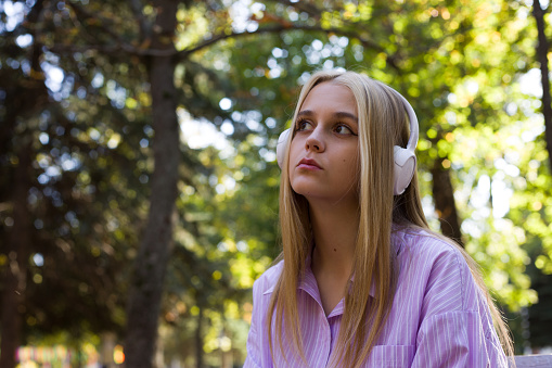 Young blonde woman in a pink shirt looks thoughtfully to the side while listening to music with white headphones on her head in a park against the background of trees