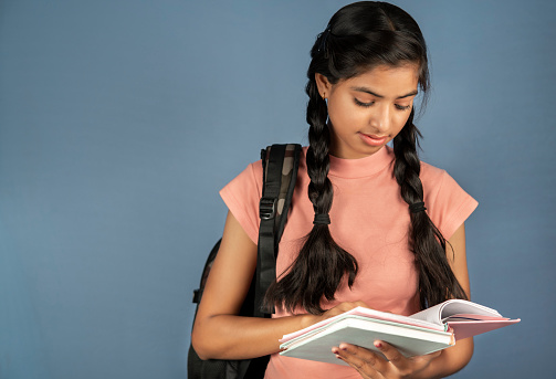 Beautiful happy female Indian student with braided hair standing against an isolated background with backpack and reading a books.