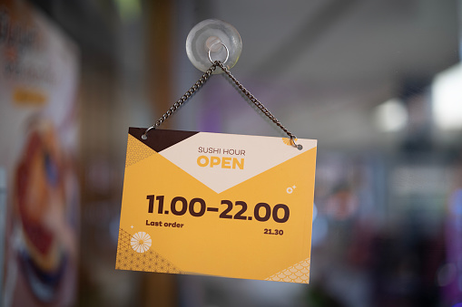 Open and welcome text sign at front door of food store with business hours, black and yellow text on creamy and yellow paper design, welcome phase for advertisement with blur inside shop menu and copy space.