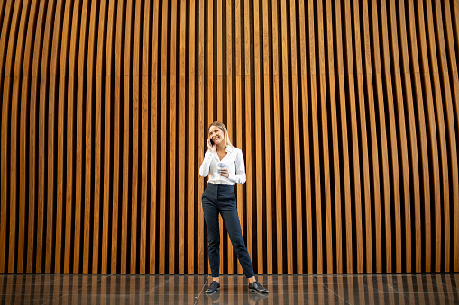 Young confident and happy businesswoman standing in office building and using mobile phone. She is wearing business casual clothes and holding reusable coffee cup