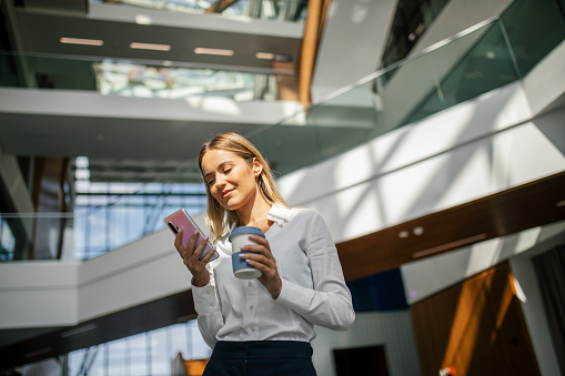 Young confident and happy businesswoman walking in office building and using mobile phone. She is wearing business casual clothes and holding reusable coffee cup