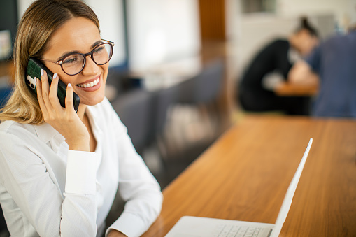 Young confident businesswoman sitting in canteen in office building and working on laptop and politely talking on mobile phone. She is wearing business casual clothes