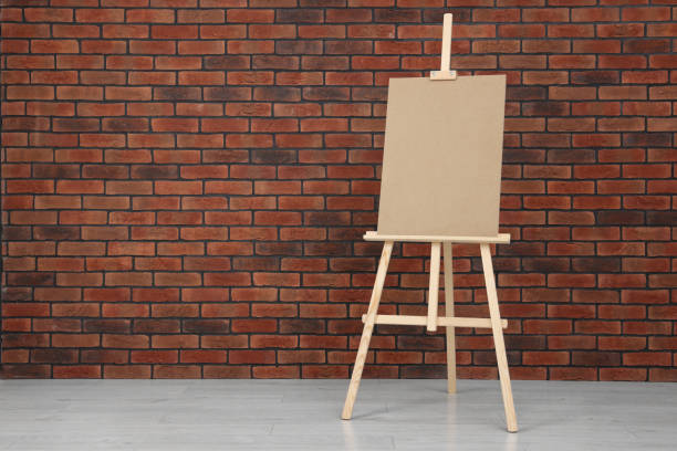 istock Wooden easel with blank board near brick wall indoors. Space for text 1721188868