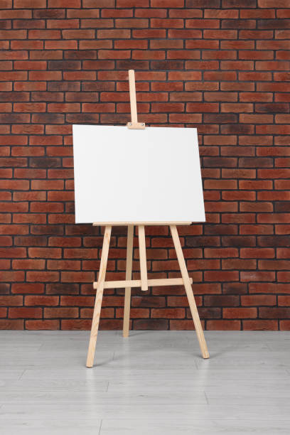 istock Wooden easel with blank canvas near brick wall indoors 1721189397
