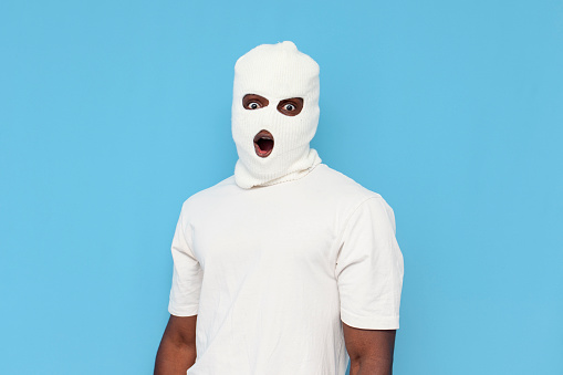 shocked african american male thug in white balaclava is surprised over blue isolated background, unknown hooligan in mask in amazement with open mouth