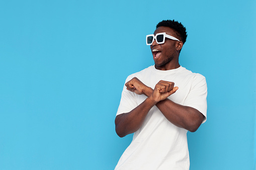 african american man in white t-shirt and sunglasses dances to music and sings on blue isolated background, man moves in disco, party concept