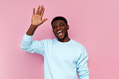 african man in blue sweater raises his hand and greets on pink isolated background, man shows empty palm and smiles