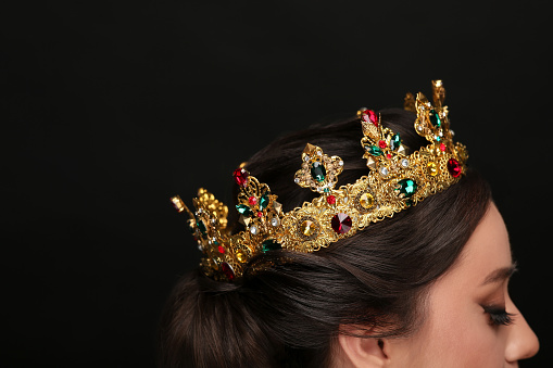 Beautiful young woman wearing luxurious crown on black background, closeup. Space for text