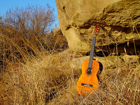 Boulder overshadows a classical guitar outdoors in dried grass as a metaphor or 'rock' music :) has an album look to scene