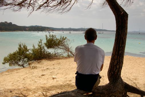 Man sitting under the tree looking at the yachts anchored in a bay of island Porquerolles, France.