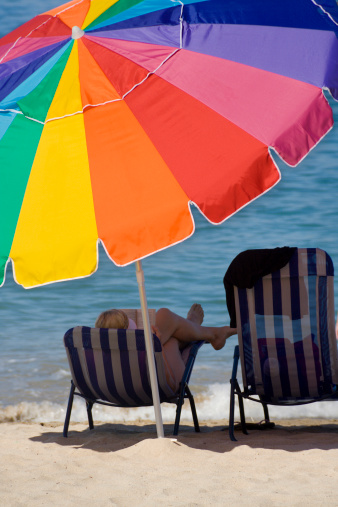 Summer leisure reading on beach chair under rainbow color umbrella sunshade at tropical climate tourist travel destination, Puerta Vallarta, Mexico. Lounge seating at water's edge, on clean white sandy beach. Vertical view of woman relaxing alone, getting away from it all and waiting for her romantic beach vacation companion. 