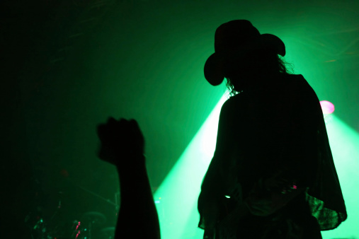 Silhouette of a guitarist on stage with a cowboy hat with fan's fist in front of green reflector