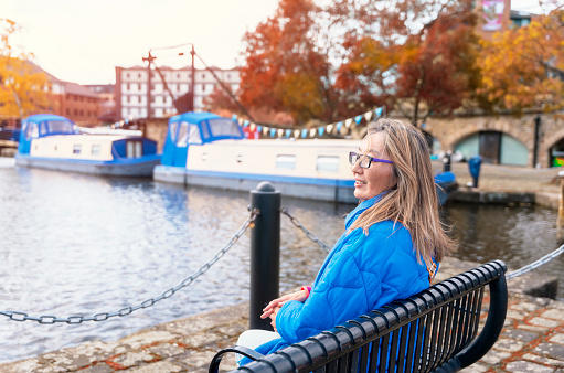 asian woman in blue jacket and glasses walking on the quays, pier, sitting on bench, looking at yacht, sailboat, boat. Lifestyle travel concept