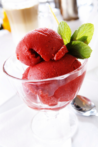 Three scoops of sweet homemade berry ice cream or refreshing sorbet of pink color decorated with juicy raspberries and fresh mint leaf served on plate on white wooden table with metal scooper utensil