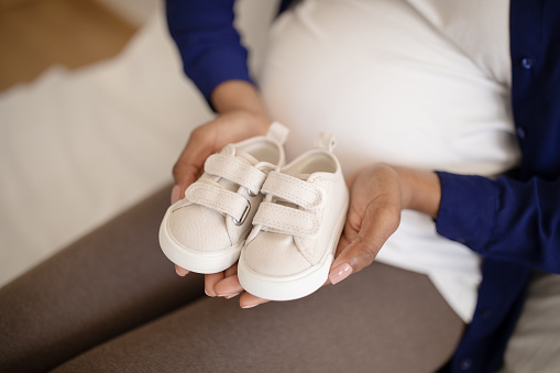 Millennial african american lady with big belly hold small shoes for future baby in room interior, cropped, close up. Childbirth, expectation of child, motherhood and family