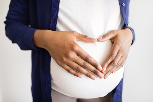 Young african american pregnant woman in casual touches heart shaped belly with hands, enjoys expecting baby on white background. Love, preparation for childbirth, motherhood and family at home