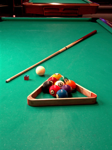 A cue stick, cue ball, chalk, rack and nine-balls on a green pool table. Colorful image.