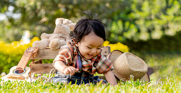 Happy baby smiling, dressed as a cowboy or farmer, sitting on the grass on a sunny day, with a rope in hand and his little guitar