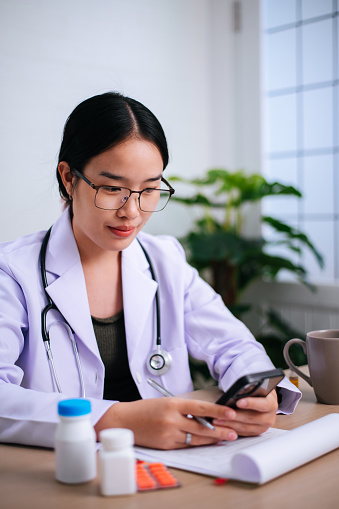 Portrait of Young Asian Female Doctor holding a prescription bottle and writing a prescription on a special form at the clinic. Healthcare, Vaccine, medical and pharmacy concepts.