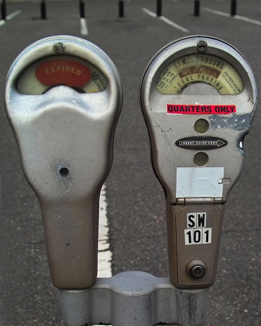 A pait of parking meters in an empty lot. They are old so the glass is dark but you can clearly read the writing behind it.