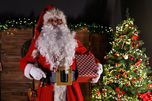 Portrait of Santa Claus standing by the glistening Christmas tree, holding his gold bell a gift box and looking at camera.
