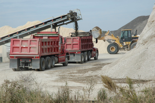 A red dump truck is loaded by a conveyor system with sifted crushed concrete for use in a freeway expansion project in east Texas as a second red dump truck is in line behind it.  A front-end loader is passing in the background and piles of the road material are all around.