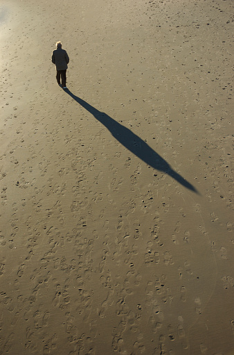 White-haired figure of senior man walking alone on the beach casting a long shadow along the sand