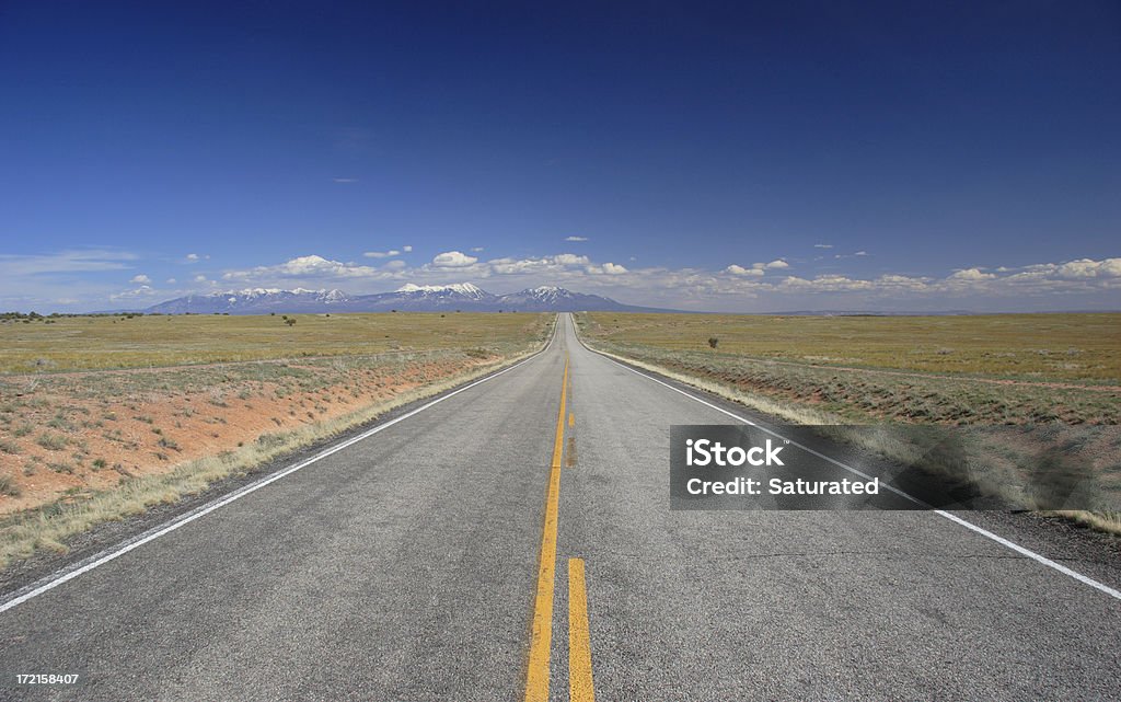 Straight Road Towards Distant Mountains A country road heads straight off into the distance towards distant mountains. Sunny day with blue sky and distant clouds over the mountains. Wide angle shot. (Utah) Asphalt Stock Photo