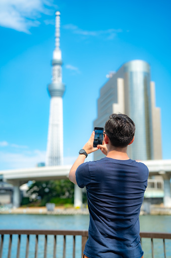 Unrecognizable young Hispanic male tourist taking photos with smartphone against broadcasting Tokyo Skytree observation tower in Japan
