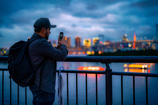 Hispanic male tourist in jacket taking photos with smarphone near Odaiba Statue of Liberty during trip in Tokyo Japan