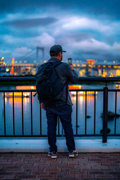 istock Back view of Hispanic unrecognizable man in casual clothes and cap with backpack standing by railing on observation deck while admiring Odaiba cityscape and river with bridge evening sky Tokyo Japan 1721615196