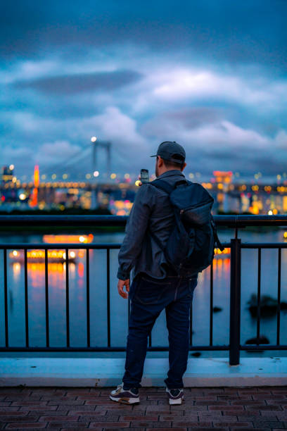 istock Back view of Hispanic unrecognizable man in casual clothes and cap with backpack standing by railing on observation deck while admiring Odaiba cityscape and river with bridge evening sky Tokyo Japan 1721615199