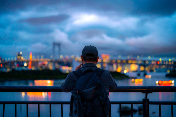 istock Back view of Hispanic unrecognizable man in casual clothes and cap with backpack standing by railing on observation deck while admiring Odaiba cityscape and river with bridge evening sky Tokyo Japan 1721615202