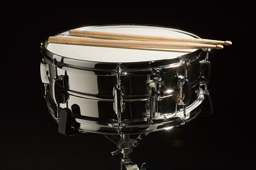 A chrome snare drum on a stand with sticks resting on the head.  Shot on a black cloth backdrop with single hard light.