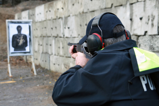 Picture of a policeman shooting Shotgun in a practice field.  The police officer is wearing his uniform and has earmuffs to protect him from the sound of the shotgun. The shooting target is blurred and visible in the background. 