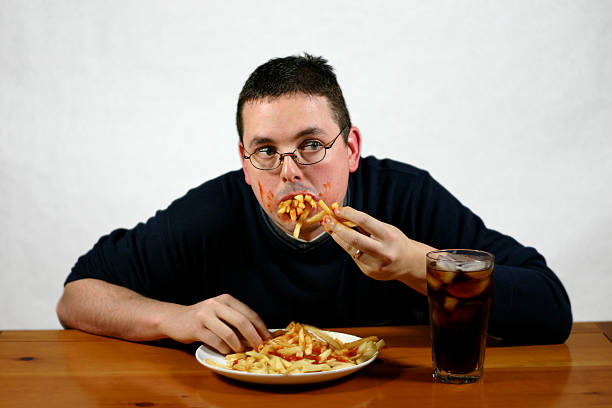 Super Size Me: Stuffed. "A man sitting at a table gorging himself with French fries soaked in catsup. Actually, his face and hands are also soaked in catsup." High Cholesterol stock pictures, royalty-free photos & images