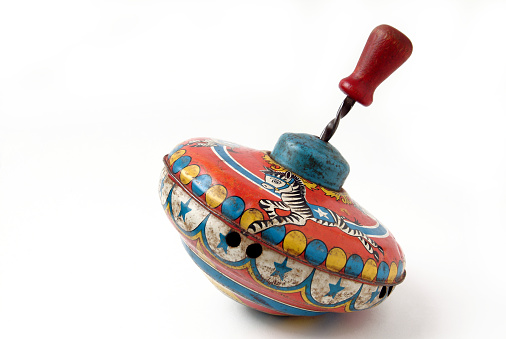 Studio image of a very old metal spinning top photographed on a white background. The top is brightly painted with a merry go round theme. The paint is very worn and the top has some dents.
